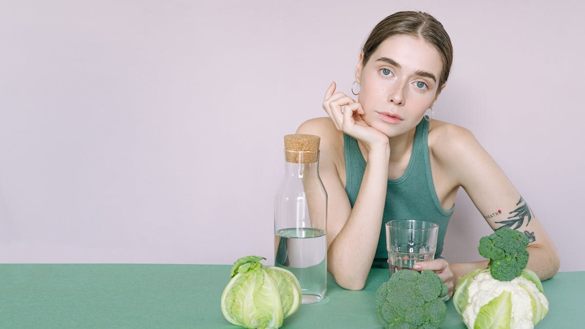 A young woman in a green top sitting with vegetables and a glass of water.